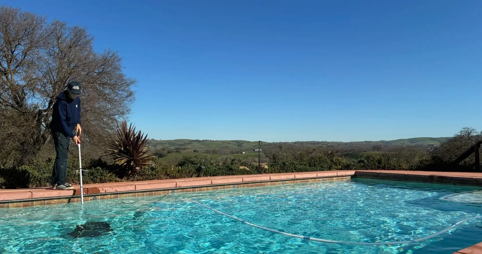 Pool Keepers technician vacuuming a backyard pool with hillside views